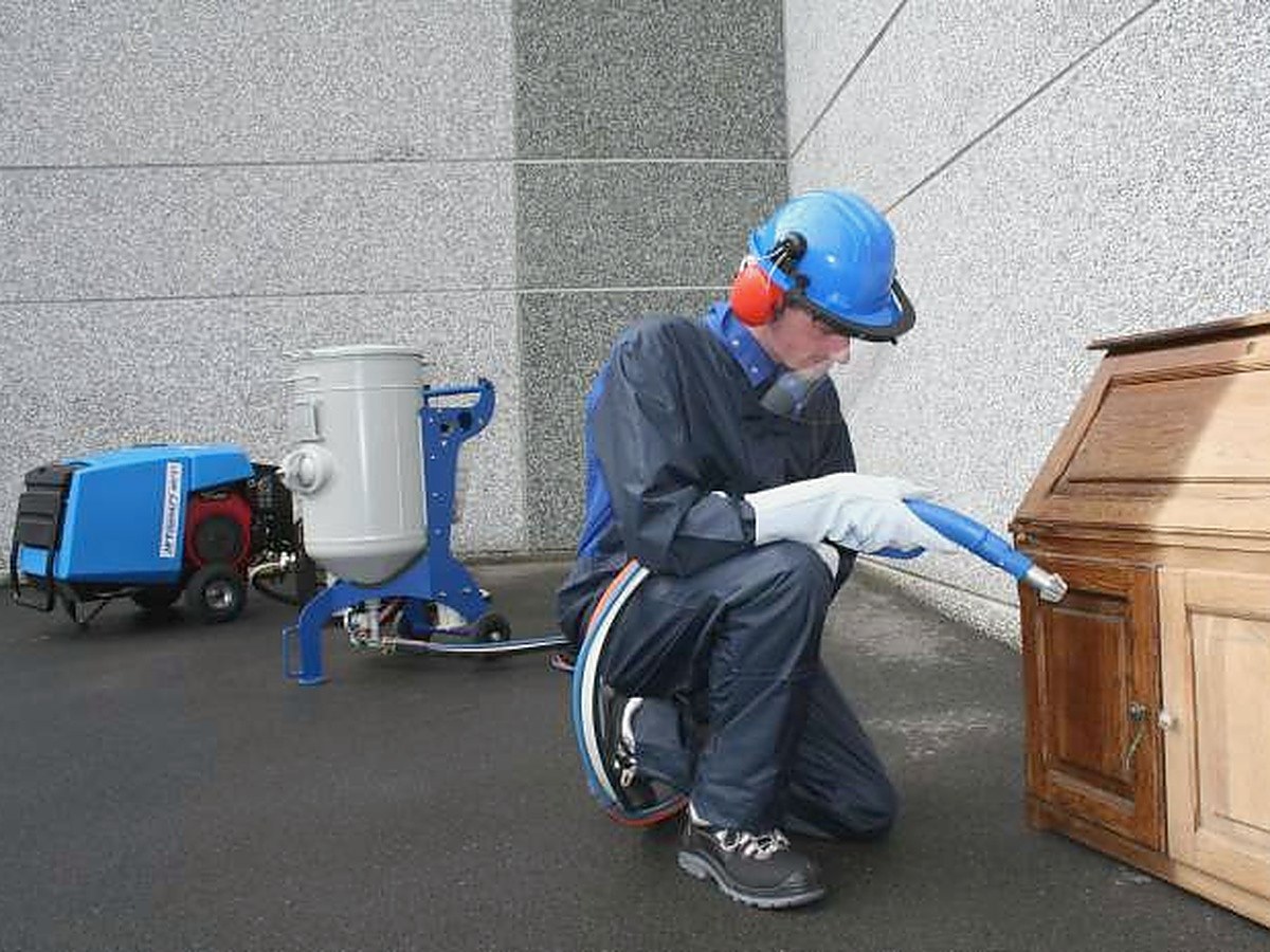 Straler die met het MCS&sup2; een houten meubel behandelt, met de straalgenerator en de compressor ernaast